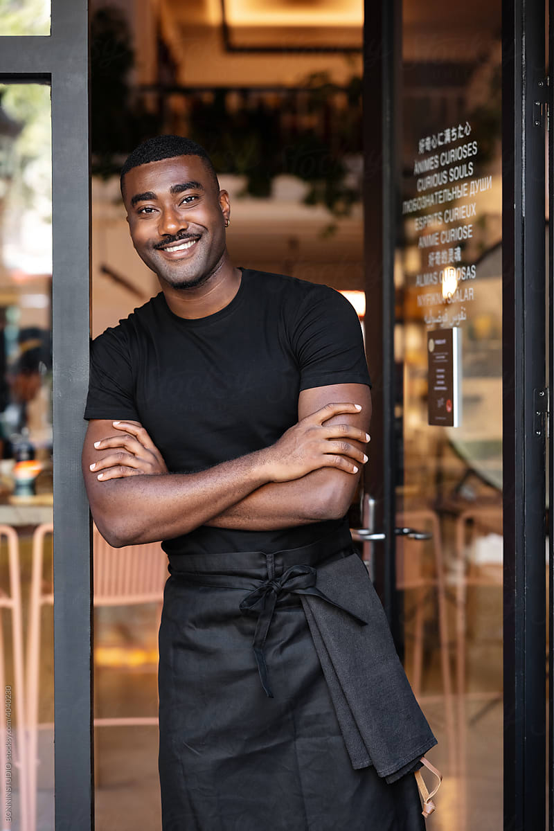 Black waiter in apron standing at doorway of cafe