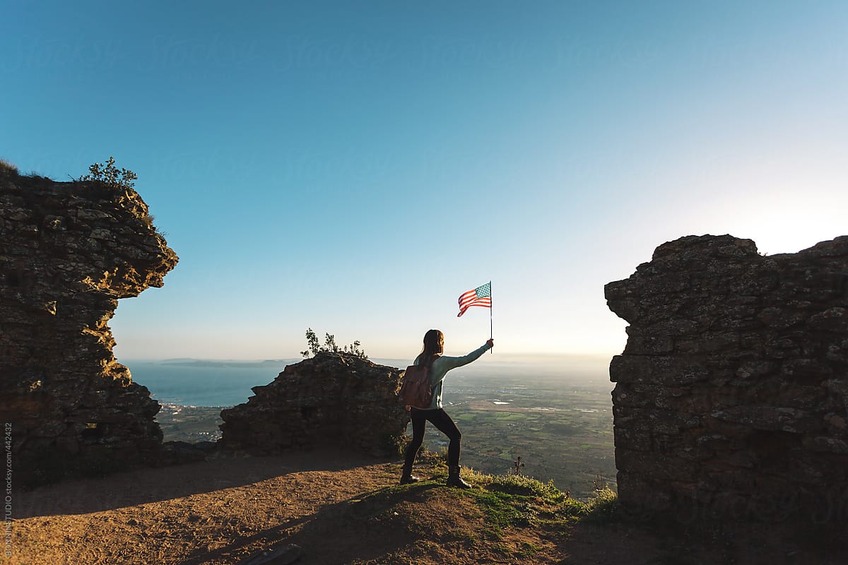 Back view of hiker woman holding an American flag on a mountain peak from far away.