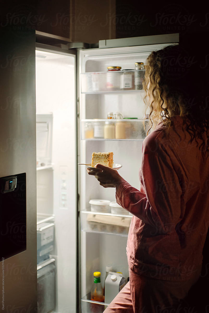 Curly-haired girl in pajamas takes a piece of cake out of the refrigerator