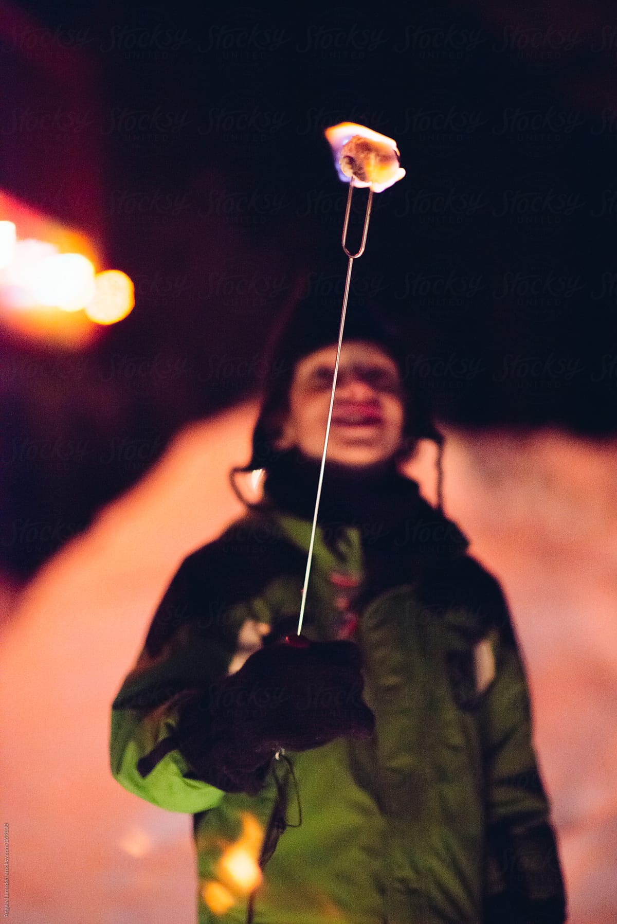 Young boy holds aloft a flaming marshmallow in the light of a camp fire in the snow at night