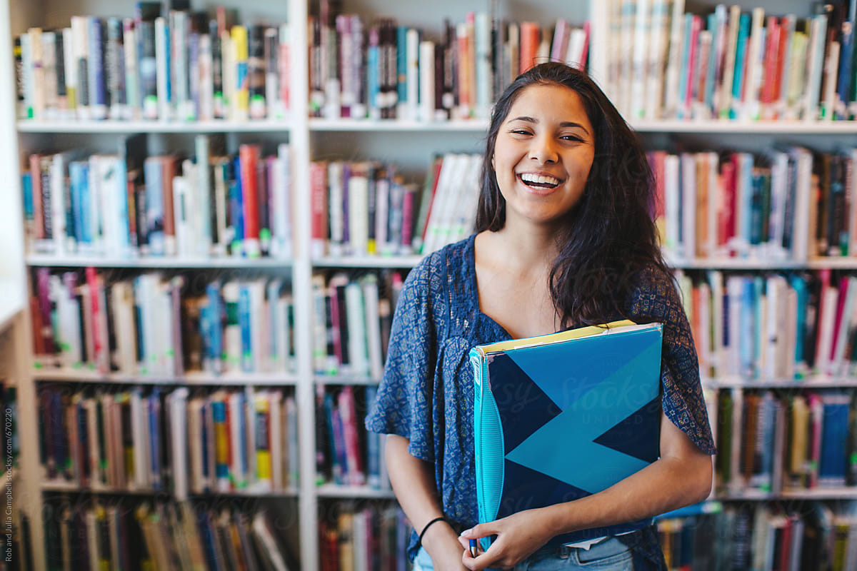 Portrait Of Happy High School Teenage Girl Holding Book Binder In Library  by Rob And Julia Campbell
