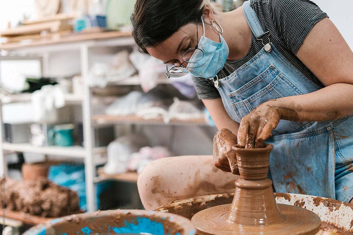 Female potter in mask crafting clay vessel in workshop