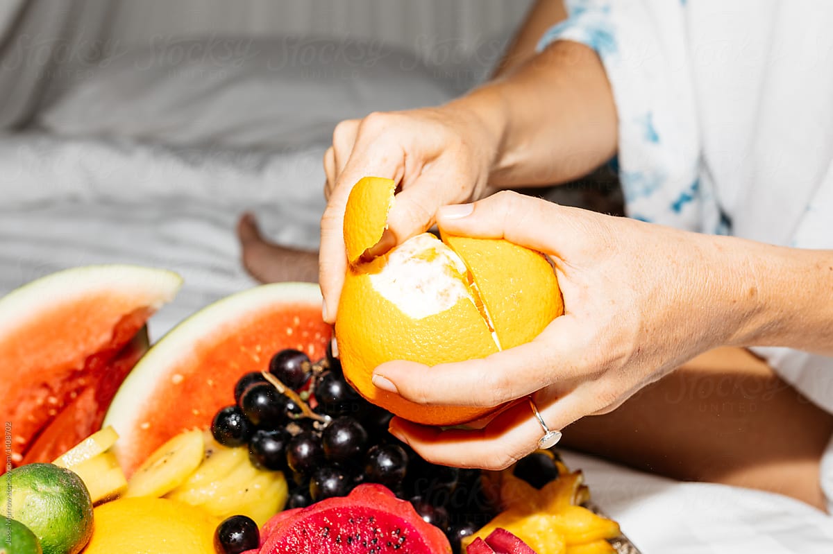 woman laying on white bed peeling and eating brightly colored orange from large tray of fruit