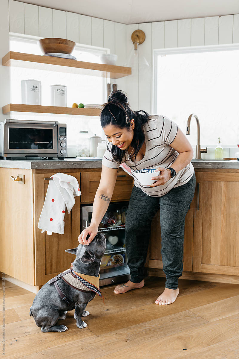 A pregnant woman cooking breakfast a small kitchen and petting her french bulldog
