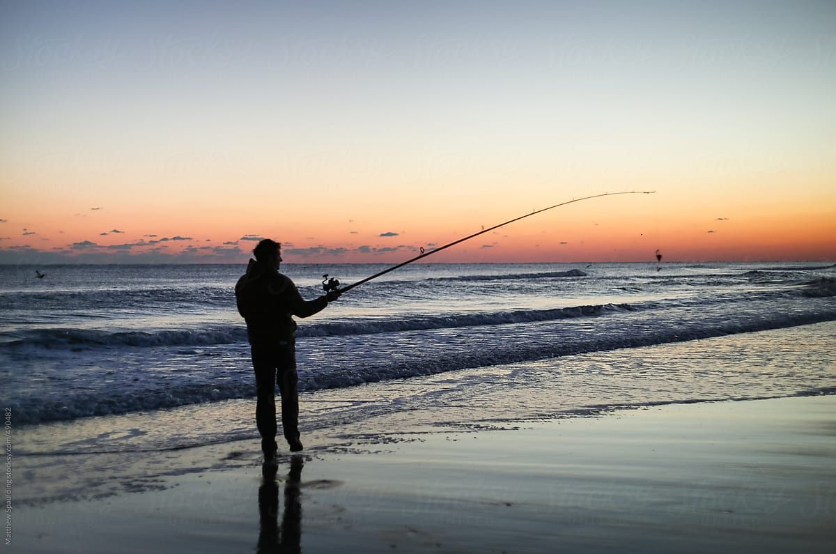 Silhouette of person surf fishing alone on ocean beach with long rod at sunset