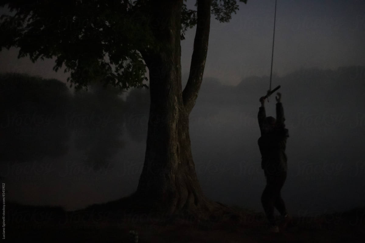 Man holding a swing at late foggy night next to a ancient tree