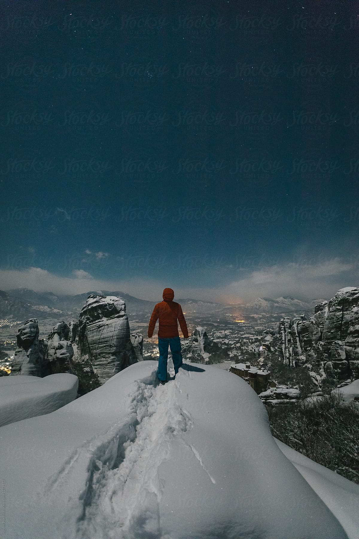 male mountaineer standing on the edge of snowcovered rocks overlooking meteora at night