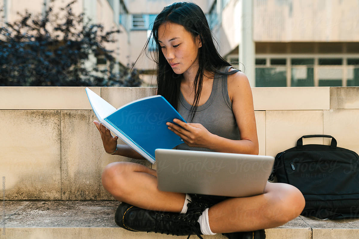 College student with laptop reading book on bench