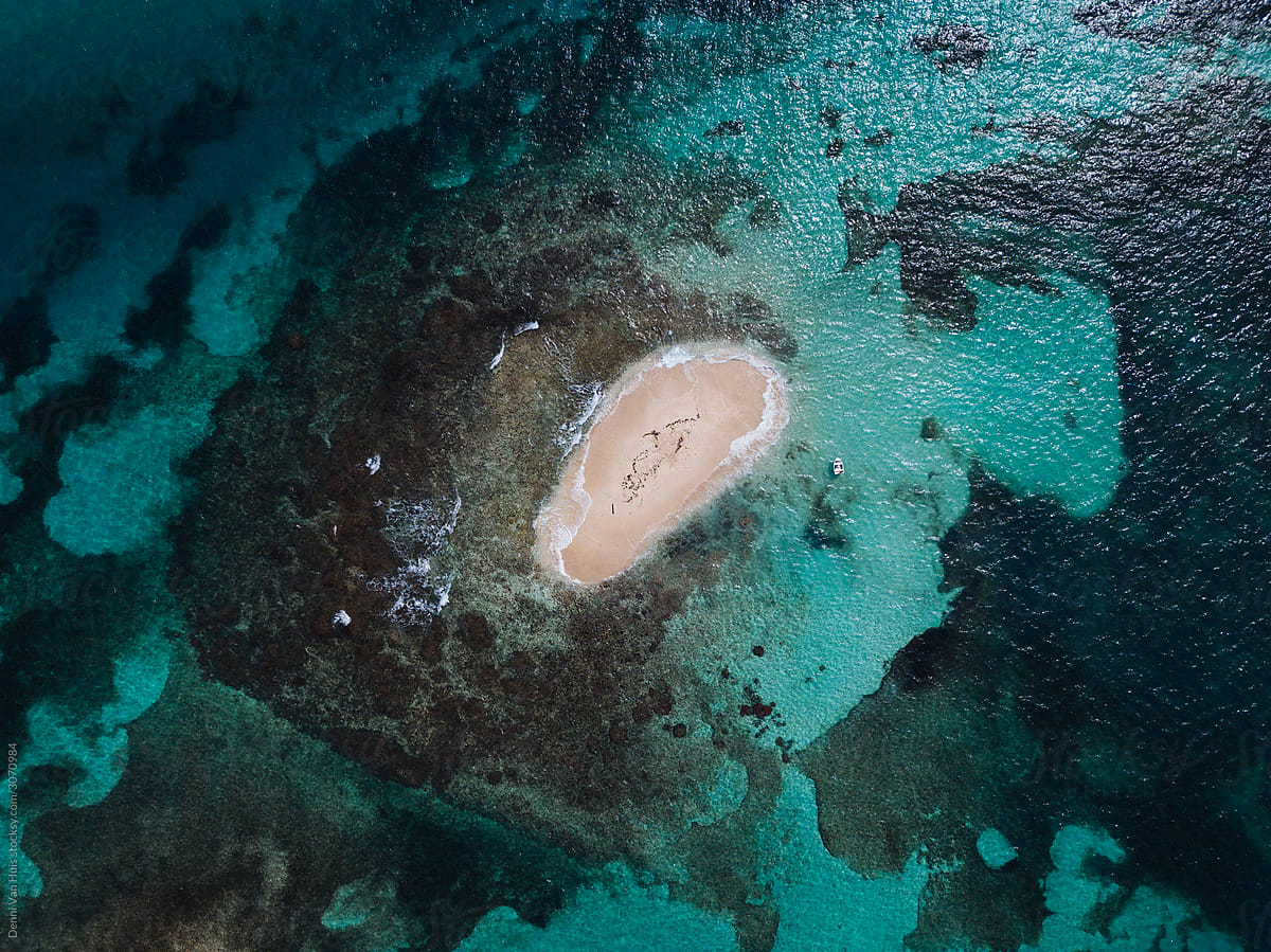 A little sandbar island in the middle of the ocean surrounded by coral and a big blue ocean.