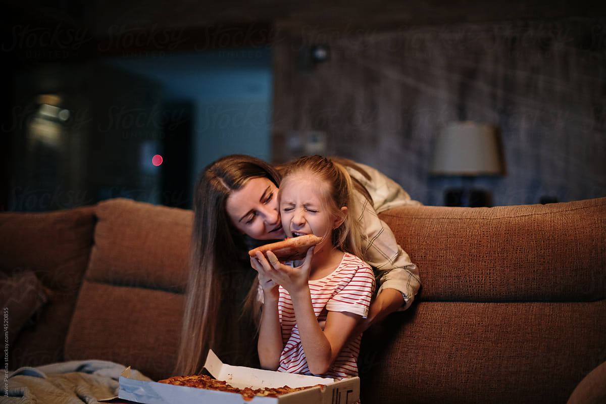 Mother teasing little girl while eating pizza on sofa
