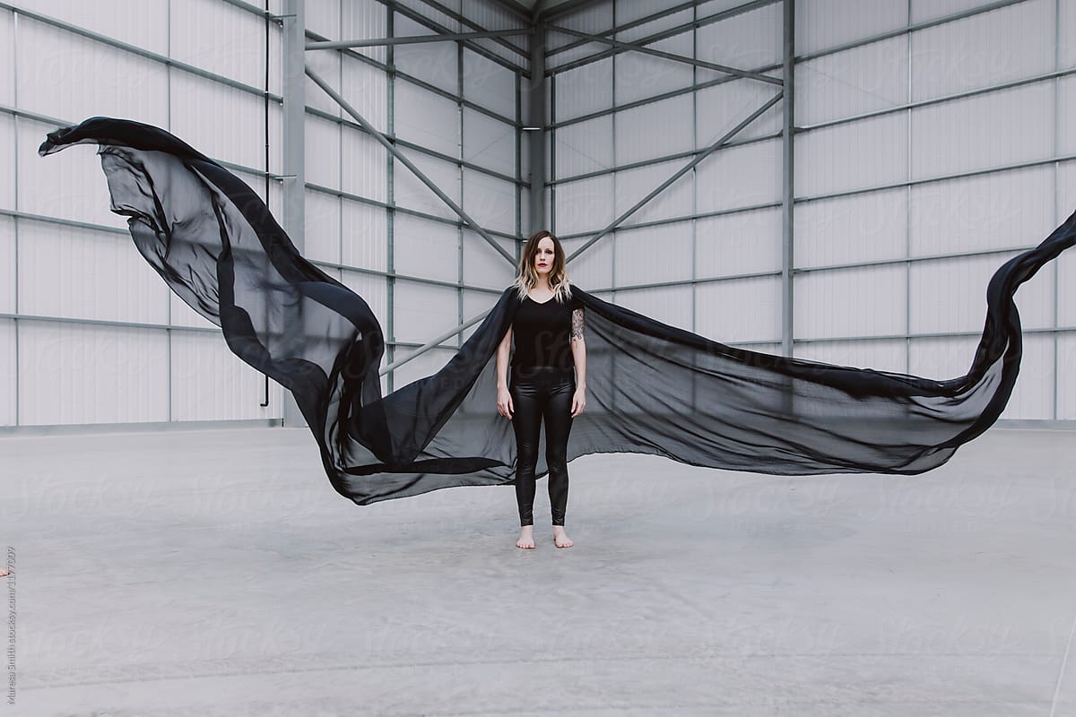 A woman wearing black standing in a warehouse. Black silk fabric that looks like wings is falling around her.