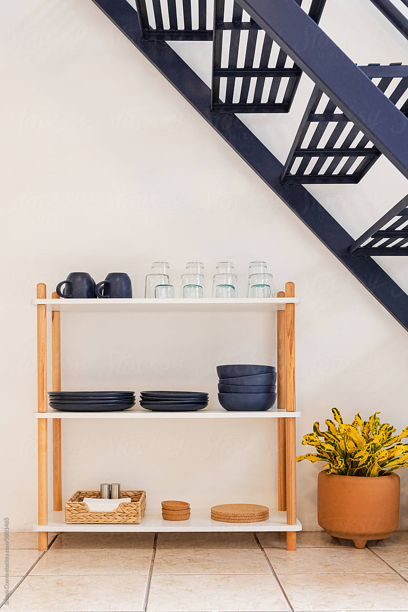 A shelf with glasses, blue ceramic, plates and bowls next to a plant