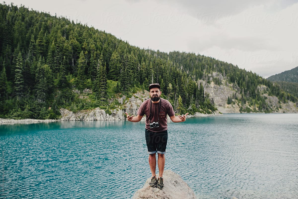 funny face of hiking bearded millennial beside blue lake