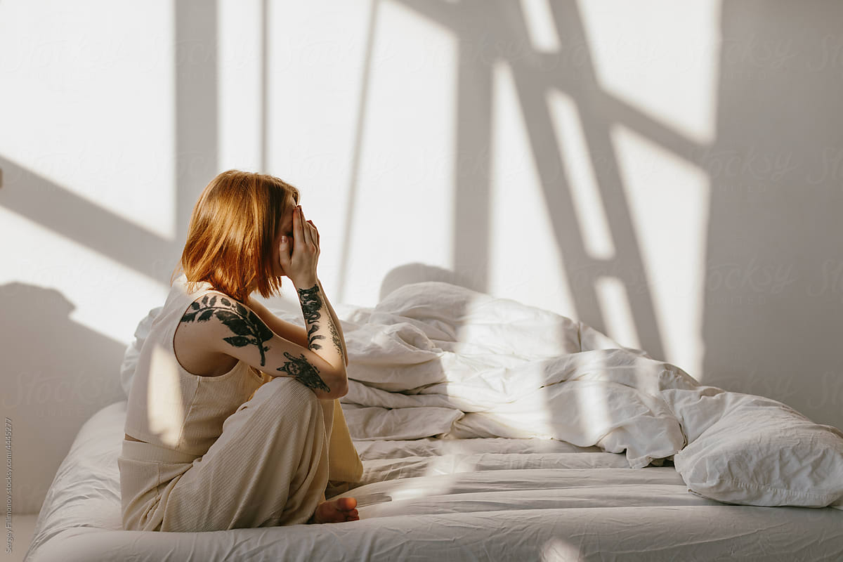 Redhead woman with headache sit on bed in sunny morning