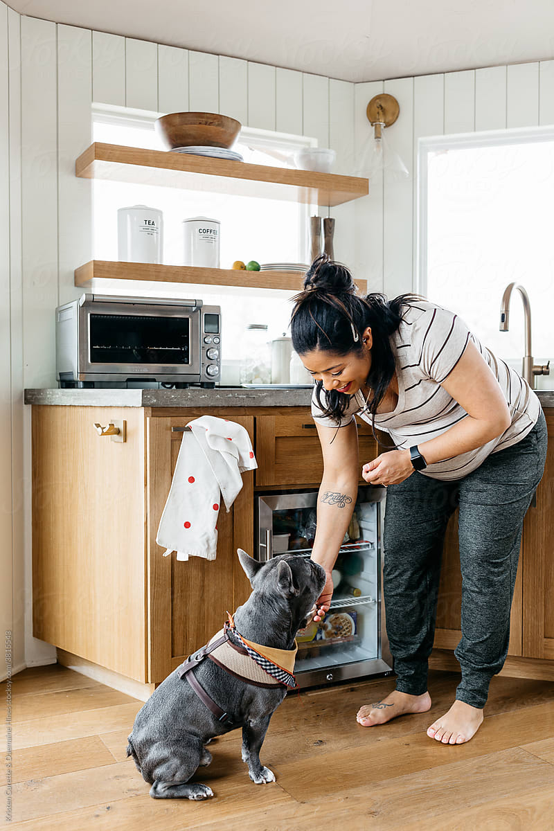A pregnant woman cooking breakfast a small kitchen and petting her french bulldog