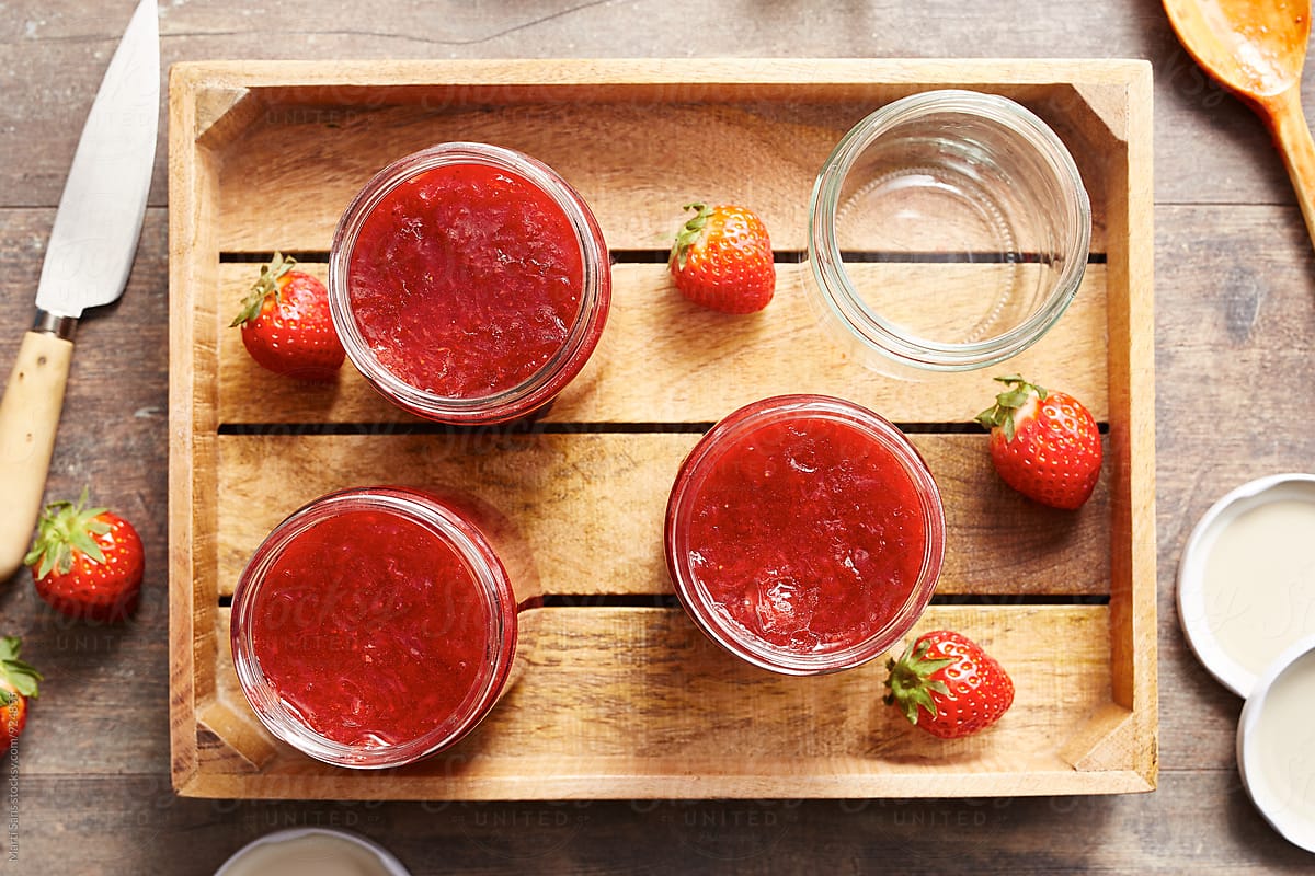 Cans full of jam with some strawberries in wooden tray