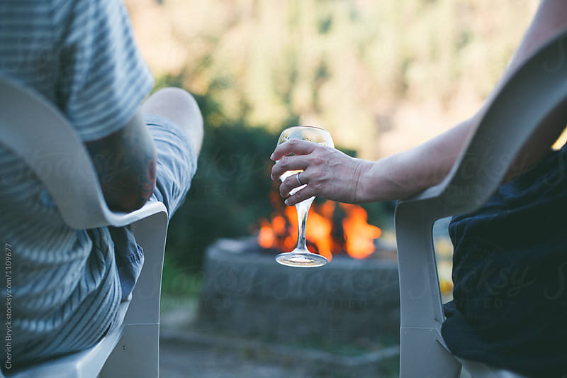 A man and woman sit in lawn chairs in front of a bonfire. The women has a wedding band and she holds a glass of wine.