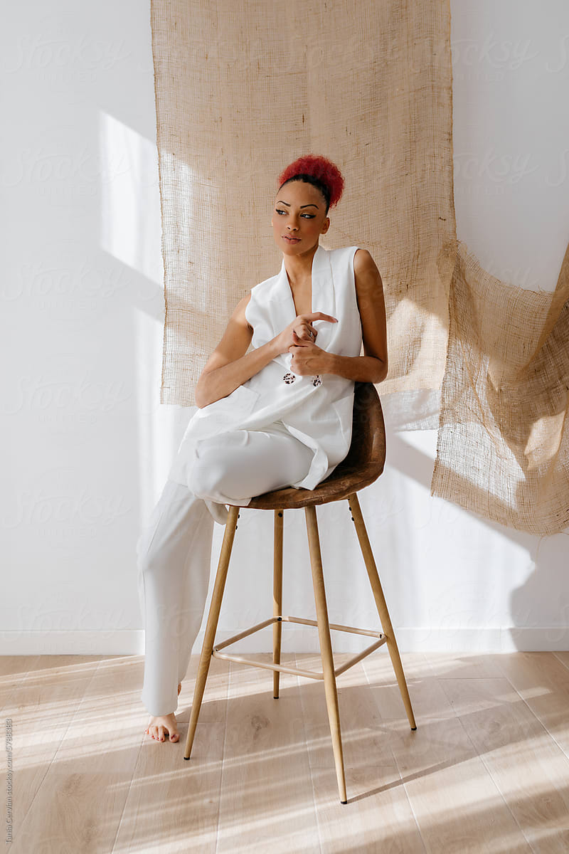 Stylish African American woman sitting on stool against jute curtain