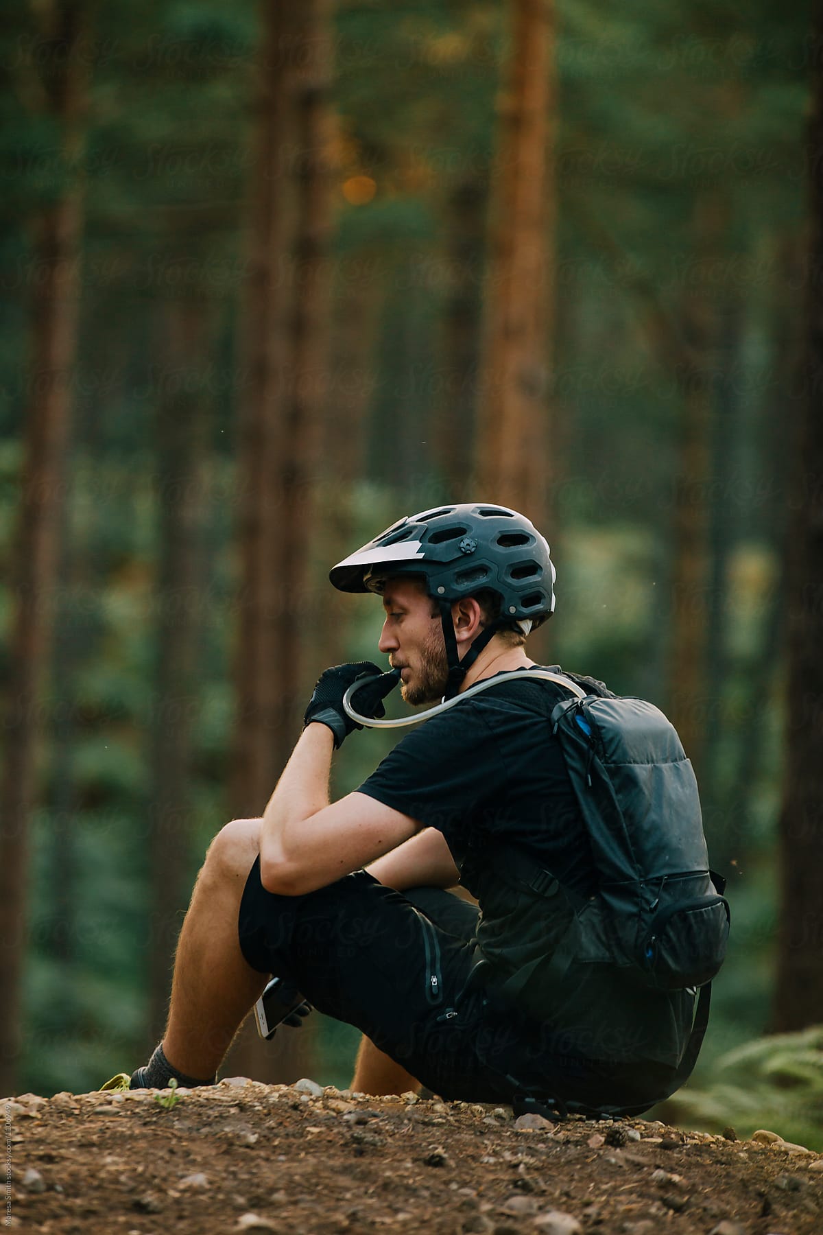 A man wearing a hydration pack takes a break to rehydrate at the top of a climb in a forest