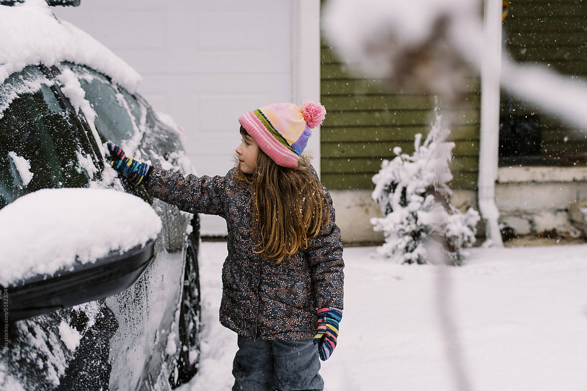 Happy toddler girl in winter gear brushing snow off car in driveway