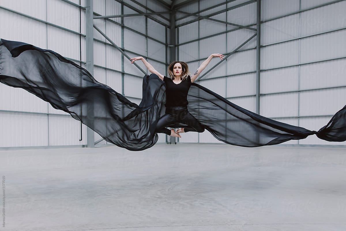 A female dancer jumping with her arms outstretched in a warehouse with black silk fabric flying either side of her