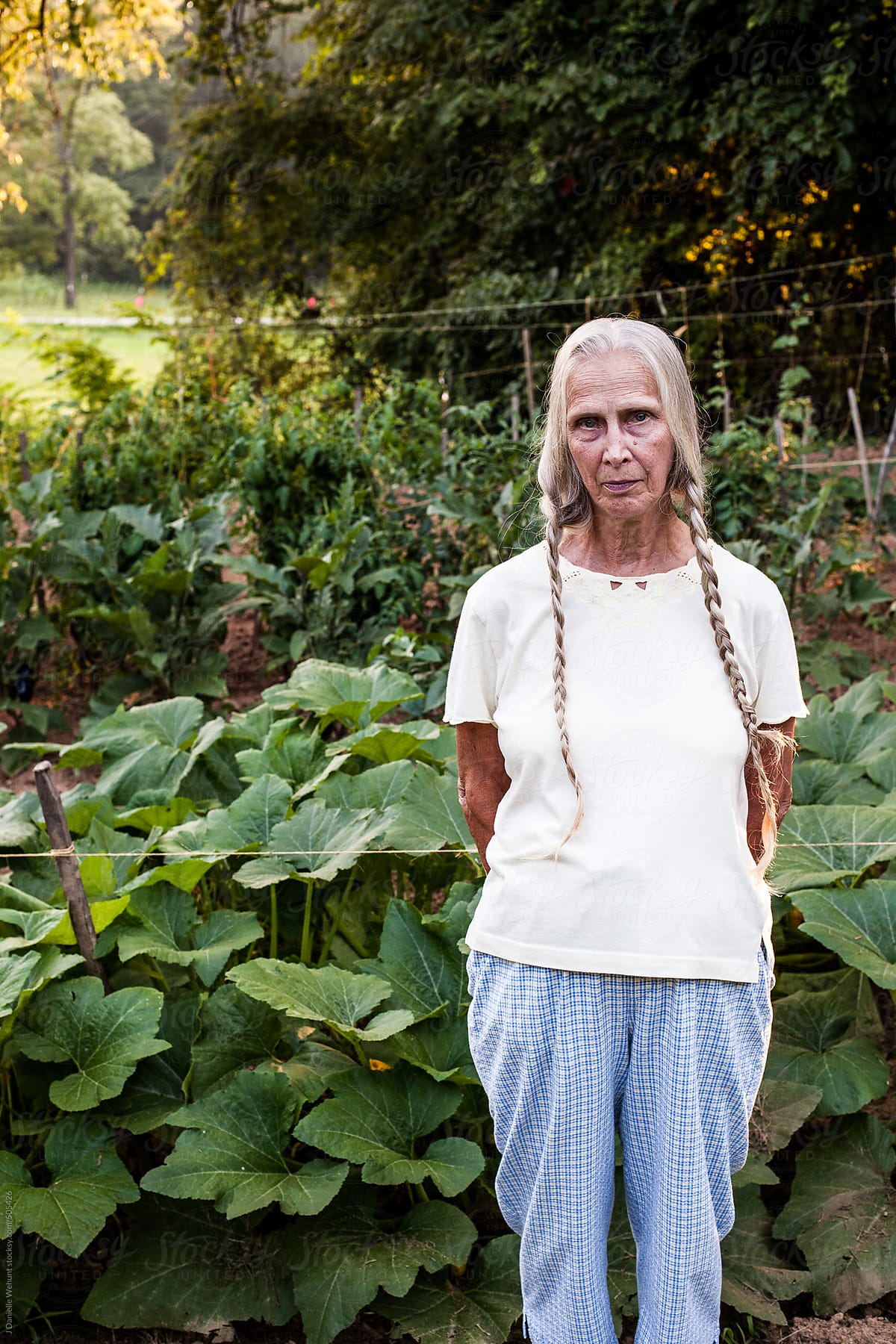 Elderly woman with blonde braids standing in front of vegetable garden