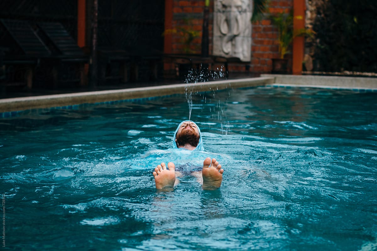Man Wearing Blue Poncho In Swimming Pool Floats On Back While Squirting Water From His Mouth