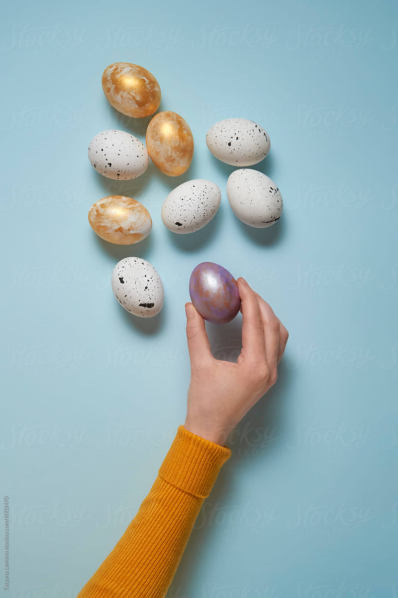 Hand in yellow sleeve decorating easter eggs on pastel blue background
