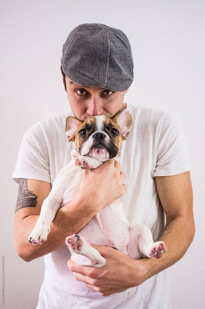 A handsome caucasian man in a hat holding and kissing a french bulldog puppy.