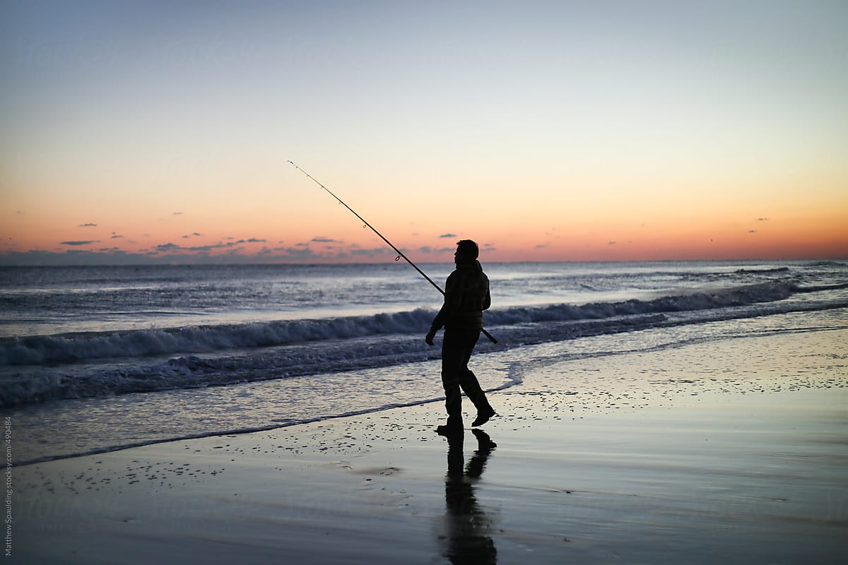 Silhouette of person surf fishing alone on ocean beach with long rod at sunset