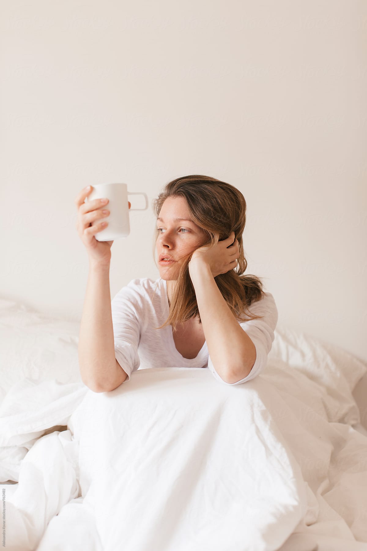 Woman In White Bed Sits And Drinks Her Morning Coffee By Stocksy