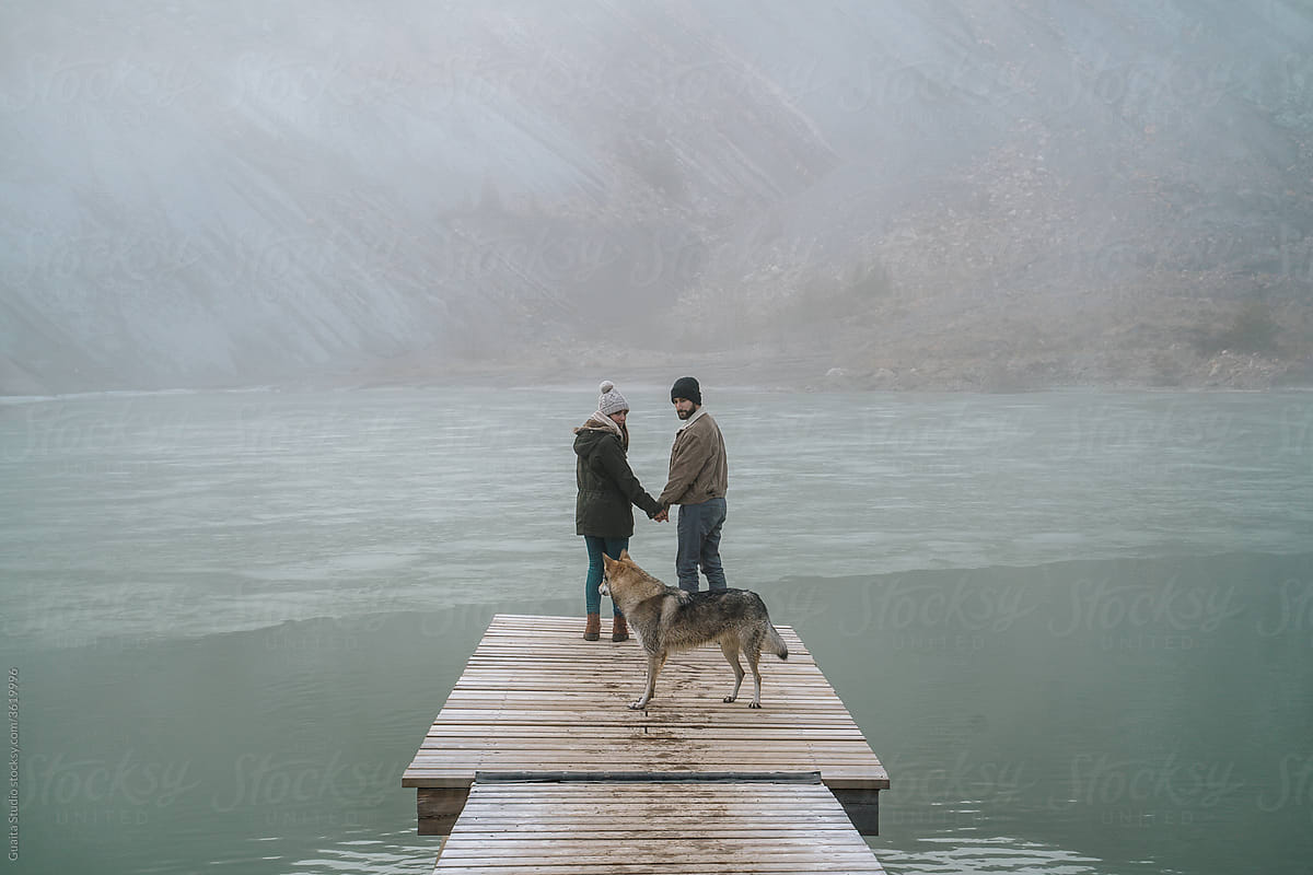 Couple on wooden dock in frozen lake in mountain with dog