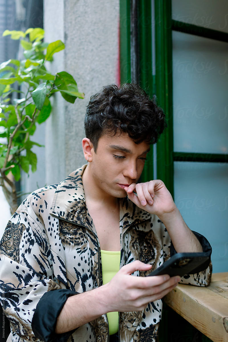 Young extravagant gay man sitting outside of a city cafeteria checking his phone