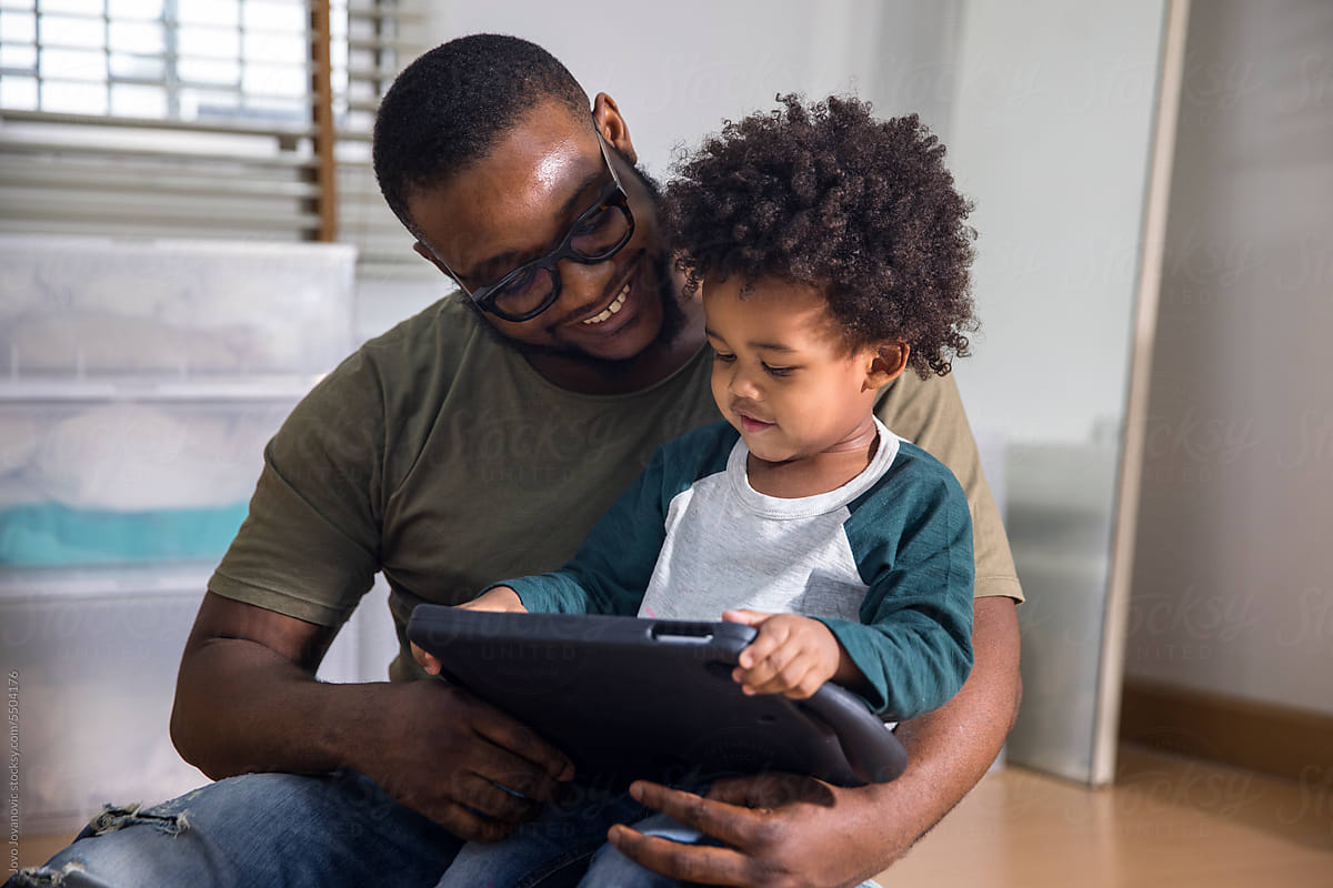 Joyful man sitting with little son using digital tablet at home