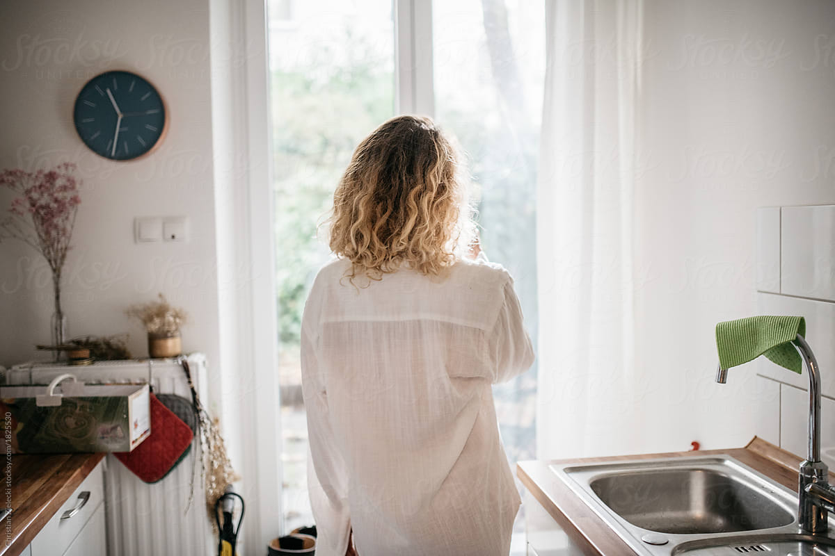 a woman in her pyjama standing in a kitchen looking out of a window