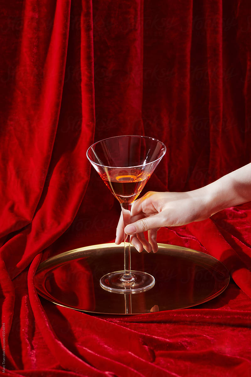 The person holds a glass of wine on red velvet curtain background