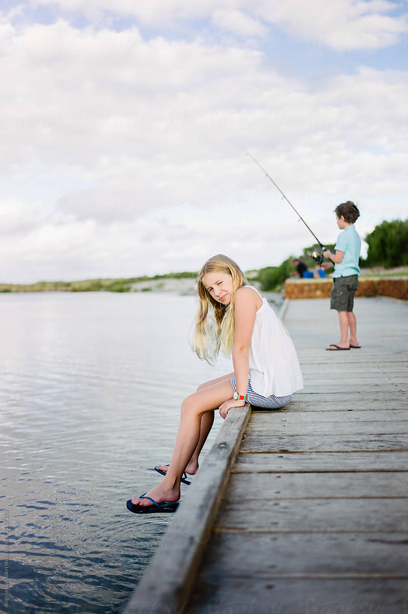Pre-teenage Girl Sitting On A Dock Beside Her Brother Fishing by