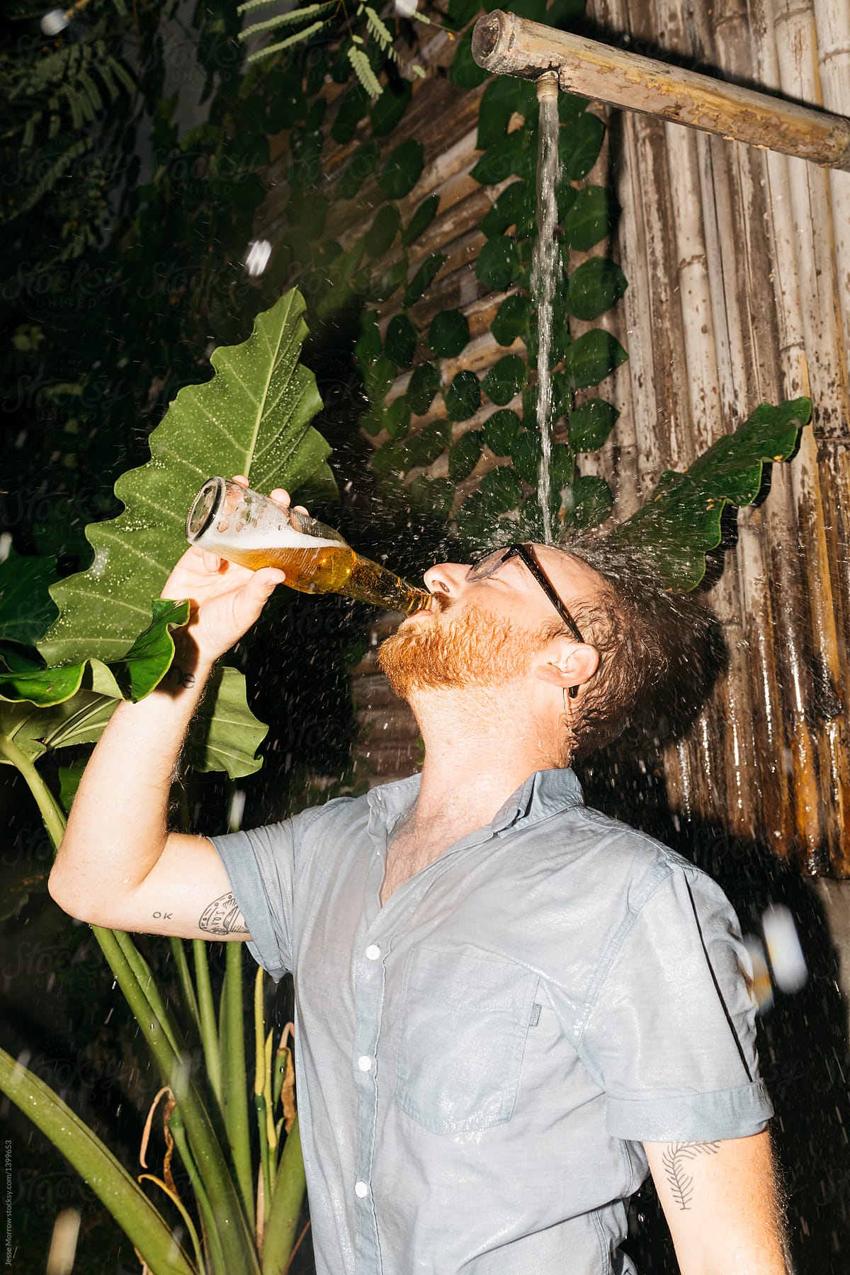 Young man drinks beer fully clothed in outdoor wild shower with large plants