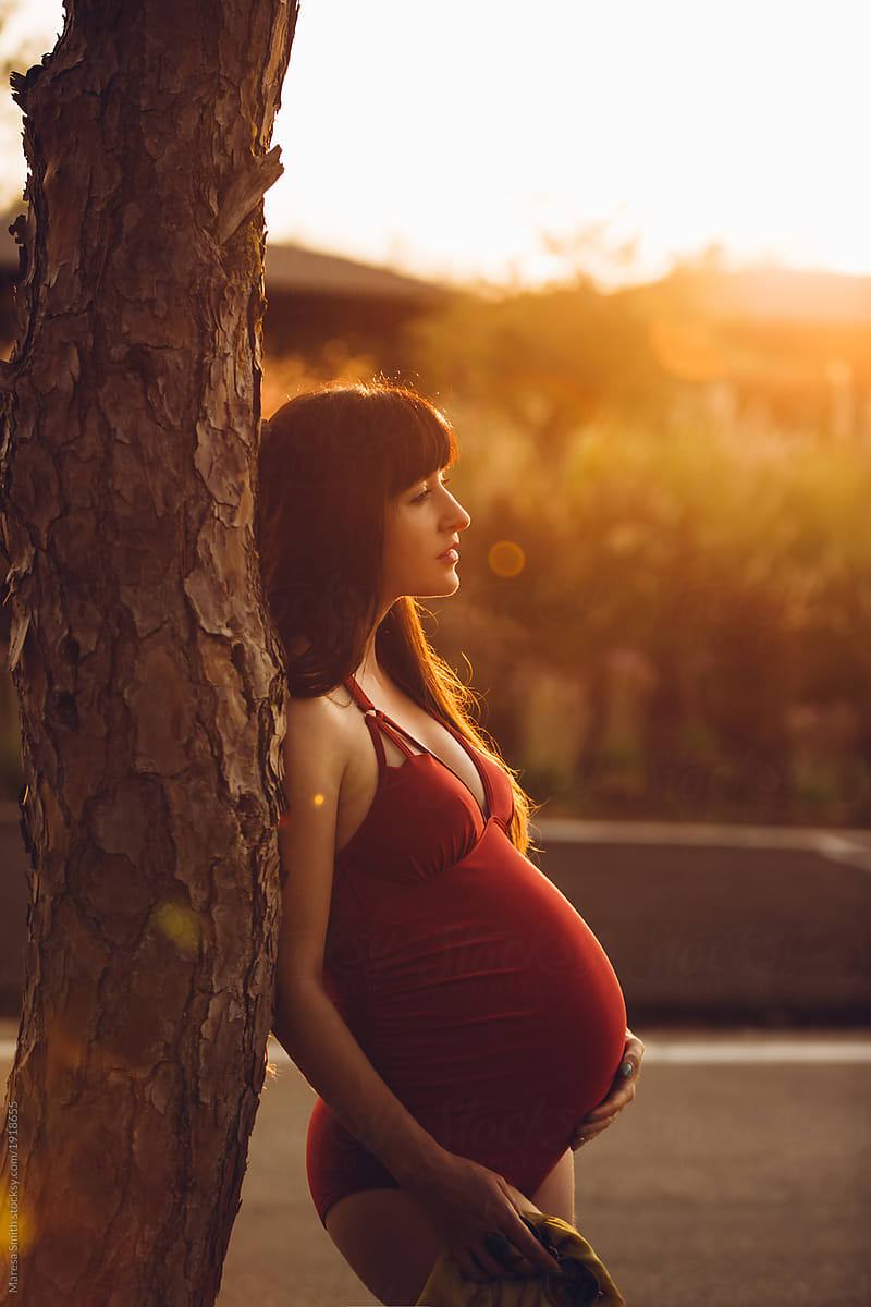 A pregnant woman in a red swimsuit at golden hour, leaning on a tree looking outward