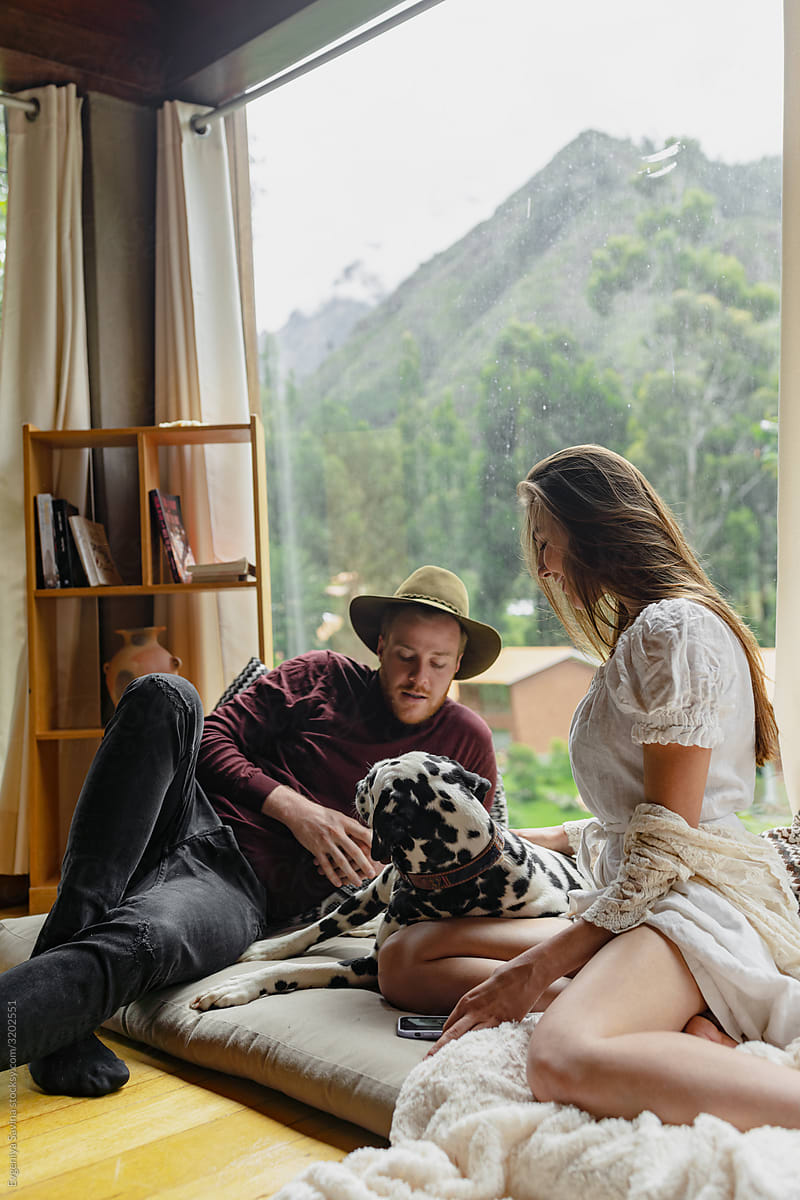 A young couple sitting in a lounge zone in a house with a Dalmatian dog and playing with it.