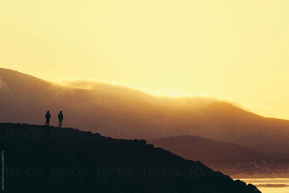 Two people stand on a hill overlooking the ocean as the sun rises