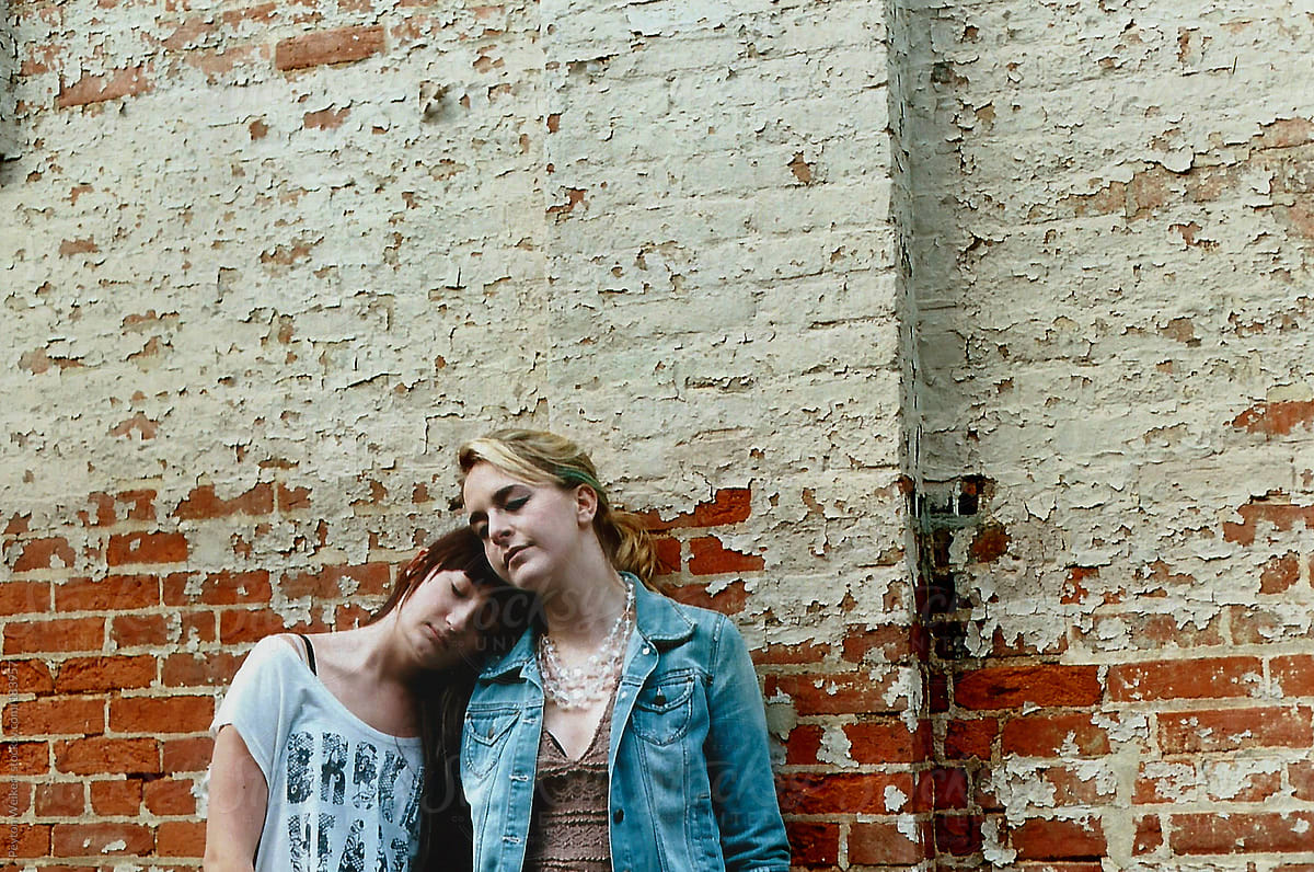 Two Intimate Girl Friends Huddle Together Against A Peeling Paint Brick Wall By Stocksy