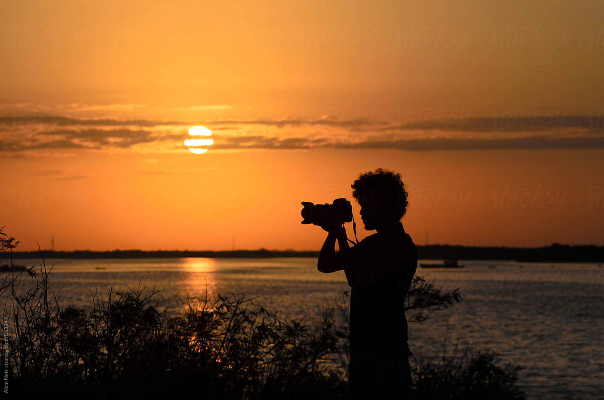 Black silhouette of photographer with his camera nearby lake at sunrise