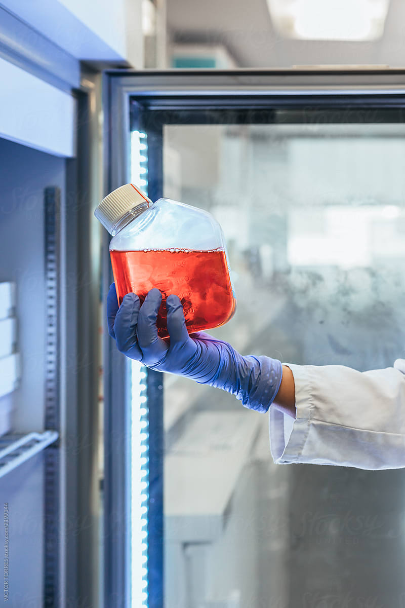Close-up biologist hand holding a plastic container with red liquid in lab.
