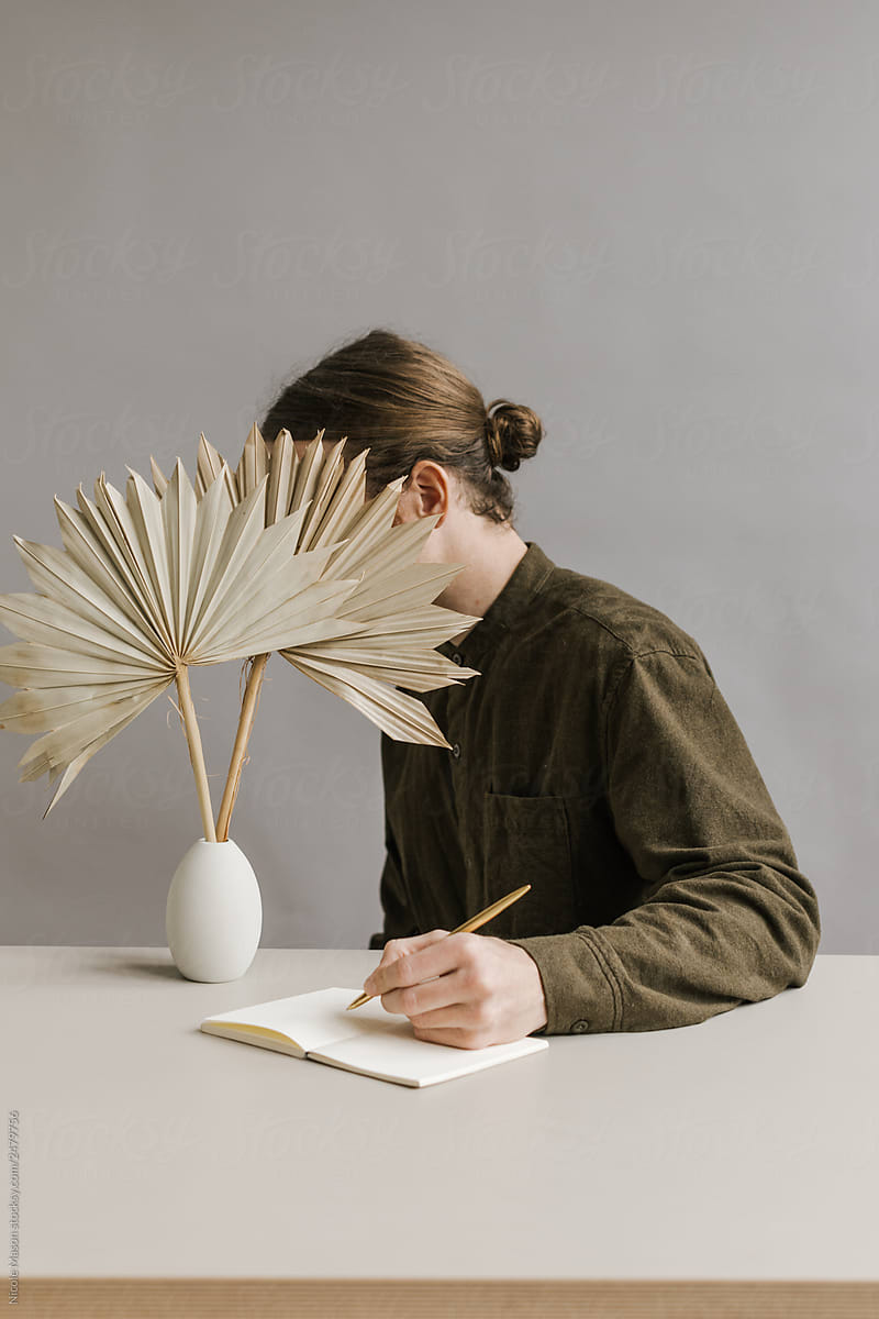 young man sitting hiding face behind dried palm leaf writing in journal