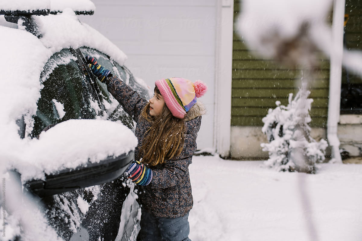 Happy toddler girl in winter gear brushing snow off car in driveway