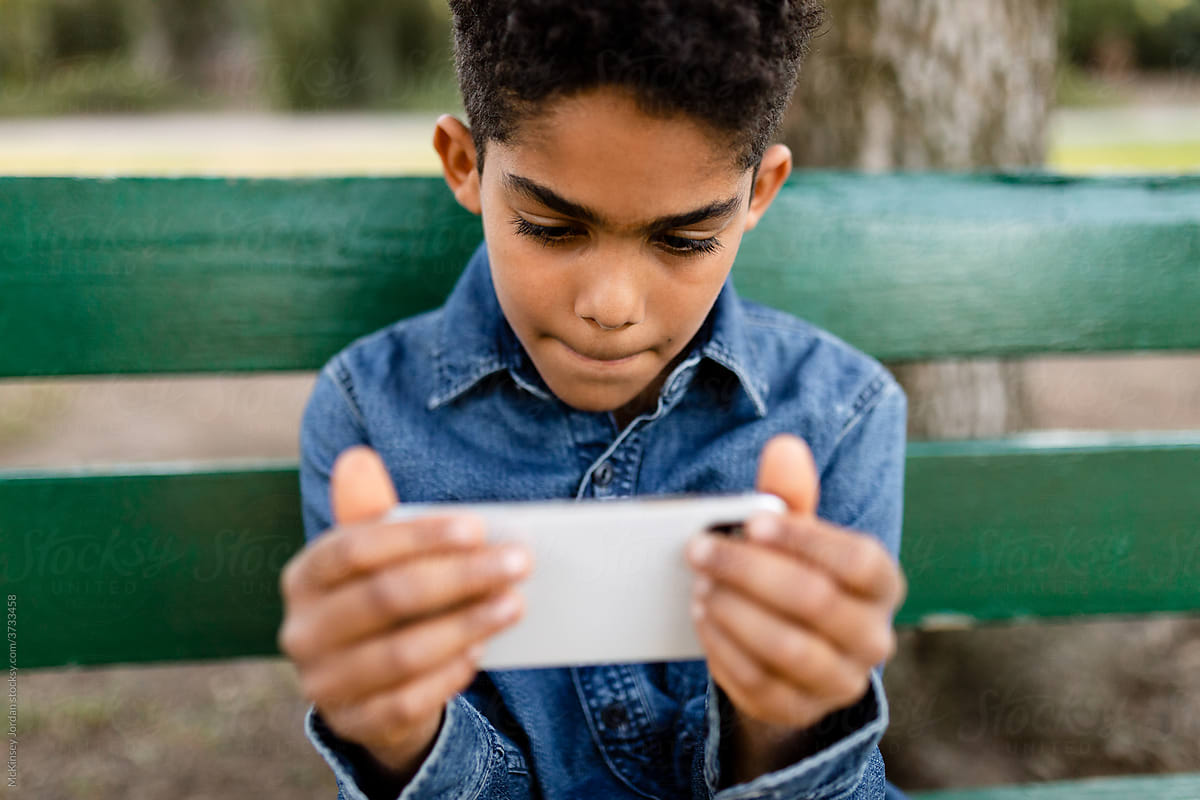 Young Boy Sits on a Park Bench and Watches a Video on His Phone by Himself