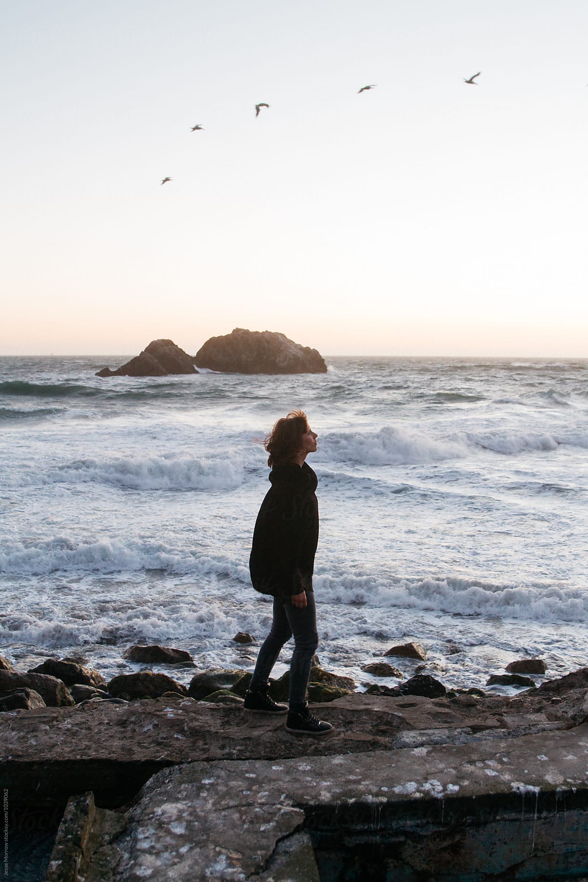 young woman walks along ocean coast during sunset while waves crash and birds fly