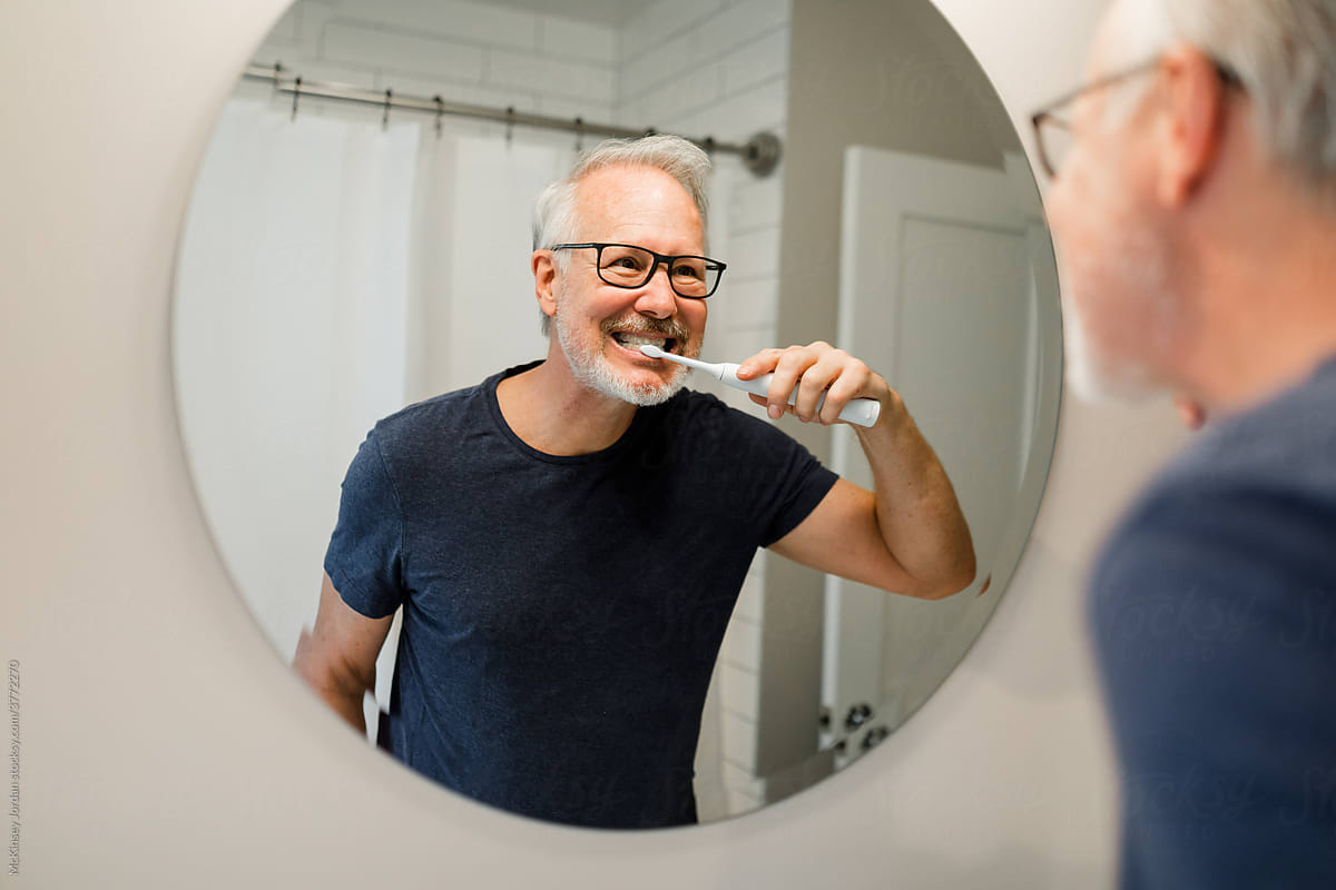 Older Gentleman Smiles While Brushing His Teeth in Bathroom Mirror