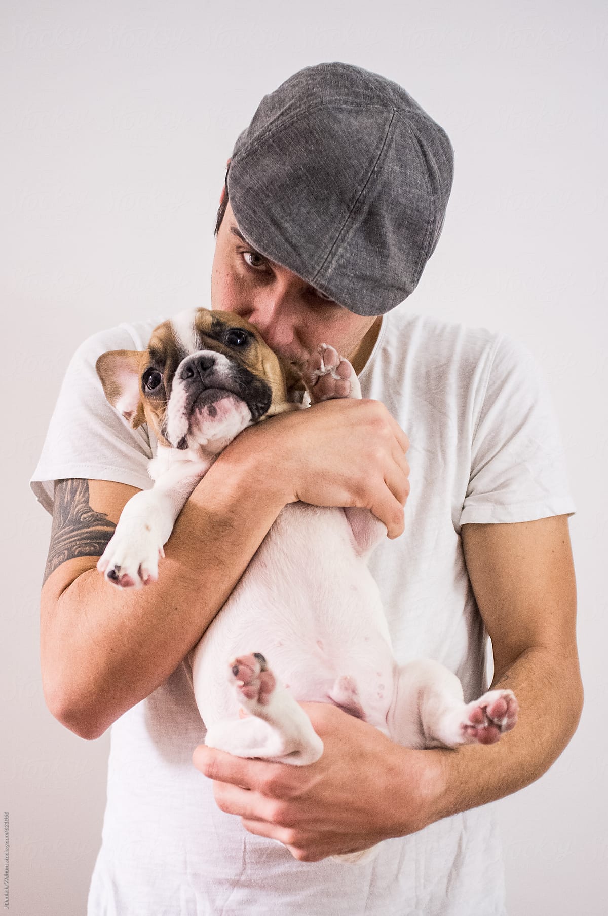 A handsome caucasian man in a hat holding and kissing a french bulldog puppy.
